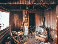 Ein verlassener, holzverkleideter Raum mit unordentlichem Boden, der mit Werkzeugen, Holzstücken und Maschinenresten übersät ist. An den Wänden sind einige Geräte und eine Elektroinstallation sichtbar. Der Raum hat zwei Türen, eine ist geöffnet. Licht fällt von außen herein.