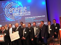 Eine Gruppe von Menschen steht auf einer Bühne bei der Preisverleihung des "Energie Start-up Bayern 2018". Sie halten einen großen Scheck und lächeln. Im Hintergrund ist ein Banner mit dem Eventnamen und Datum zu sehen.