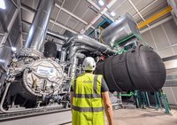 Ein Arbeiter in Warnweste und Helm steht vor einer großen, industriellen Maschine mit zahlreichen Rohren und einem großen schwarzen Zylinder. Die Maschine scheint Teil einer Anlage zur Energieerzeugung oder -verteilung zu sein, möglicherweise von einem Unternehmen namens Johnson Controls.
