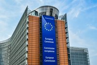 Das Bild zeigt die Fassade der Europäischen Kommission in Brüssel. An der Wand ist ein großes Banner mit dem EU-Sternenlogo und der Aufschrift „Europese Commissie“, „Commission européenne“ und „European Commission“ angebracht. Der Himmel ist blau und bewölkt.