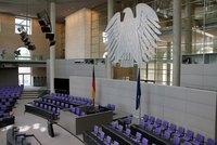 Das Bild zeigt den Innenraum des Deutschen Bundestages mit einem großen Bundesadler über der Rednertribüne. Der Raum ist minimalistisch gestaltet, mit lila Stühlen und einer modernen Architektur. An den Wänden hängen Flaggen von Deutschland und der Europäischen Union.