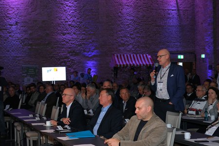 Ein Mann mit Brille und Anzug steht auf und spricht zu einem Publikum auf einer Veranstaltung. Die Zuschauer sitzen an Tischen und hören aufmerksam zu. Die Raumbeleuchtung ist in Lila gehalten.