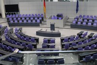 Ein Blick auf den Plenarsaal mit vielen lila Sitzen, die in einem Bogen angeordnet sind. In der Mitte steht ein Rednerpult vor einer Erhöhung. Im Hintergrund sind Symbole der deutschen und europäischen Flaggen. Der Raum wirkt modern und funktional.