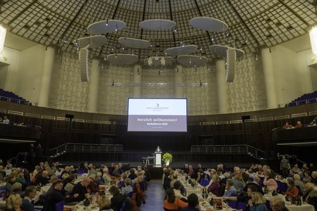 Eine große Veranstaltung in einem Saal mit vielen Teilnehmern, die an Tischen sitzen. Auf einer Bühne steht ein Redner, und ein Bildschirm zeigt die Begrüßung „Herzlich willkommen!“. Die Stimmung ist festlich und die Anordnung der Plätze deutet auf ein gemeinsames Essen hin.