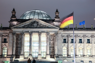 Das Bild zeigt das deutsche Bundeshaus, den Reichstag, bei Nacht. Im Vordergrund weht die deutsche Flagge, während die EU-Flagge daneben sichtbar ist. Die beeindruckende Fassade des Gebäudes ist beleuchtet und die Kuppel ist deutlich zu erkennen.