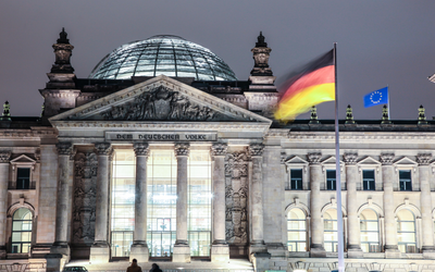 Das Bild zeigt das deutsche Bundeshaus, den Reichstag, bei Nacht. Im Vordergrund weht die deutsche Flagge, während die EU-Flagge daneben sichtbar ist. Die beeindruckende Fassade des Gebäudes ist beleuchtet und die Kuppel ist deutlich zu erkennen.