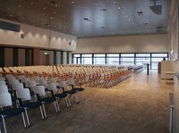 Ein großer, heller Konferenzraum mit mehreren Reihen von Stühlen, die ordentlich aufgestellt sind. Die Stühle sind überwiegend in Weiß und haben schwarze Sitzpolster. Große Fenster lassen Tageslicht herein und bieten einen Blick nach draußen. Der Raum ist bereit für eine Veranstaltung.