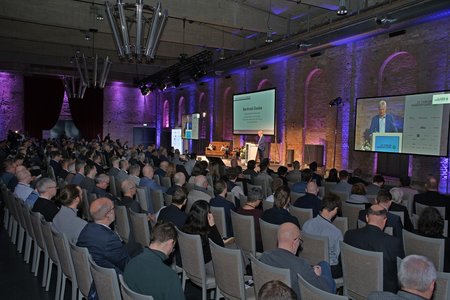 Eine große Menschenmenge sitzt in einem Konferenzsaal, während ein Redner auf einer Bühne spricht. Im Hintergrund sind zwei große Bildschirme mit Präsentationsinhalten sichtbar. Die Atmosphäre ist formal und professionell.