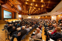 Eine große Menschenmenge sitzt in einem modernen Konferenzsaal. Eine Präsentation wird an eine Leinwand projiziert, während eine Person auf der Bühne spricht. Der Raum hat eine attraktive Holzausstattung und ist gut beleuchtet.