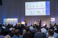 Eine Konferenzszene mit einem Publikum, das einem Vortrag lauscht. Eine Dozentin präsentiert an einem Bildschirm, der den Brauereiprozess zeigt. Zwei Referenten stehen an Tischen, während das Publikum aus Männern und Frauen besteht, die aufmerksam zuhören.