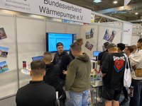 Eine Gruppe von Menschen steht an einem Stand des Bundesverbands Wärmepumpe e.V. Einige schauen auf einen Bildschirm und diskutieren. Auf dem Tisch sind verschiedene Informationsmaterialien und Getränke. Der Stand hat eine helle, moderne Gestaltung und ist mit Bildern dekoriert.