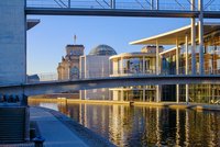Das Bild zeigt eine moderne architektonische Szene mit einem Wasserweg und einer überbrückenden Konstruktion. Im Hintergrund ist das Reichstagsgebäude mit seiner charakteristischen Kuppel und der Flagge Deutschlands zu sehen, umgeben von klaren Himmel und warmem Licht bei Sonnenaufgang.
