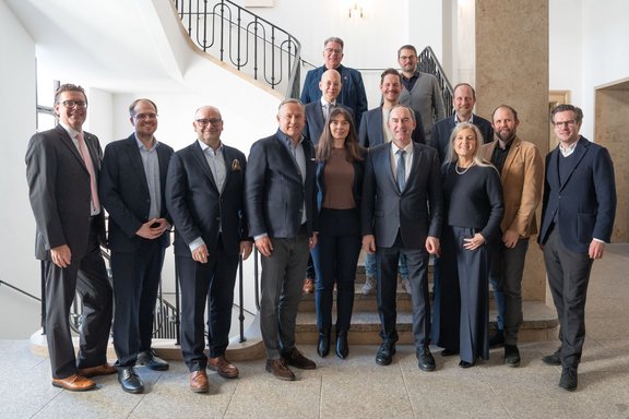 Eine Gruppe von 15 Personen steht im Innenbereich auf einer Treppe. Sie tragen formelle Kleidung und posieren lächelnd für das Foto. Die Umgebung ist hell und modern, mit klaren Linien und einer offenen Atmosphäre.