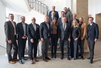 Eine Gruppe von 15 Personen steht im Innenbereich auf einer Treppe. Sie tragen formelle Kleidung und posieren lächelnd für das Foto. Die Umgebung ist hell und modern, mit klaren Linien und einer offenen Atmosphäre.