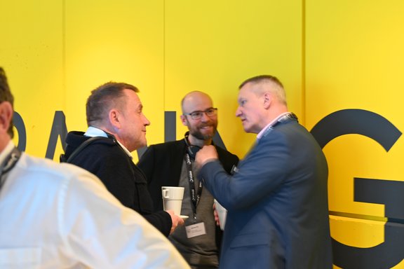 Drei Männer stehen in einem informellen Gespräch vor einer gelben Wand mit dem Wort "DACH". Sie halten Kaffeebecher und wirken engagiert.