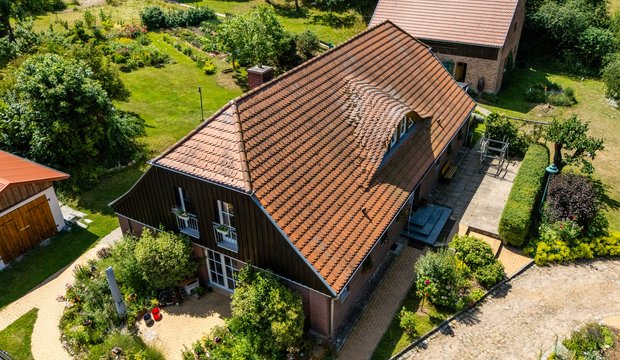 Luftaufnahme eines ländlichen Hauses mit roten Ziegeldächern. Das Gebäude ist von einem gepflegten Garten mit Blumen und Bäumen umgeben. Im Hintergrund sind Wiesen und Felder sichtbar, die die idyllische Umgebung betonen. Das Grundstück ist mit einem gepflasterten Weg und einer Einfahrt ausgestattet.