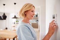 Eine Frau mit kurzen, blonden Haaren stellt einen Thermostat an der Wand ein. Sie trägt ein hellblaues Oberteil und schaut konzentriert auf das Gerät. Im Hintergrund ist eine moderne Küche zu erkennen.