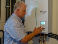 Eine Person stellt ein Bedienfeld eines Heizgeräts ein. Das Gerät zeigt eine Temperatur von 34°C an. Im Hintergrund sind Rohre sichtbar, die mit dem Heizsystem verbunden sind. Die Szene vermittelt den Eindruck technischer Wartung und Überwachung.