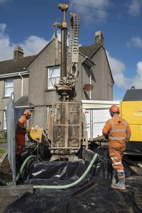 Zwei Arbeiter in oranger Arbeitskleidung stehen neben einem Bohrgerät auf einer Baustelle vor einem Haus. Das Bohrgerät ist in einer schwarzen Folie eingebettet, und grüne Schläuche führen davon weg. Der Himmel ist blau mit einigen Wolken.