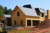Ein unvollendetes Holzhaus steht auf einer Baustelle. Die Wände sind aus Sperrholz und das Dach ist mit schwarzem Material gedeckt. Um das Gebäude herum liegen Baumaterialien, Holz und Erde. Im Hintergrund sind Bäume und ein blauer Himmel sichtbar.