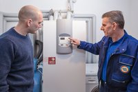 Ein Techniker zeigt einem Mann die Steuerungseinheit eines Heizgeräts. Der Techniker zeigt mit dem Finger auf die Anzeige, während der Mann neugierig zuhört. Im Hintergrund sind Rohre und weitere Geräte sichtbar.