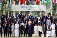 Eine Gruppe von internationalen Regierungsvertretern steht auf einer Treppe, umgeben von Flaggen verschiedener Länder. Im Vordergrund sind mehrere Personen in formeller Kleidung, die für ein Gruppenfoto während einer Klimakonferenz posieren. Das Banner oben zeigt "COP28 UAE" und das Jahr "2023".