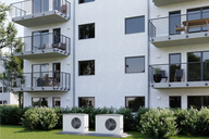Zwei Klimaanlagen stehen im Vordergrund eines modernen Wohngebäudes mit drei Stockwerken. Balkone sind mit Stühlen und Tischen möbliert. Die Fassade ist hell, und es gibt zahlreiche Fenster, durch die das Tageslicht strömt. Grüne Sträucher umgeben die Basis des Gebäudes.