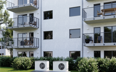 Zwei Klimaanlagen stehen im Vordergrund eines modernen Wohngebäudes mit drei Stockwerken. Balkone sind mit Stühlen und Tischen möbliert. Die Fassade ist hell, und es gibt zahlreiche Fenster, durch die das Tageslicht strömt. Grüne Sträucher umgeben die Basis des Gebäudes.