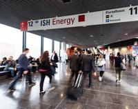 Ein belebter Hallenbereich einer Messe mit Menschen, die sich bewegen. Am oberen Bildrand ist ein Schild mit der Aufschrift „12 ISH Energy“ sichtbar. Im Hintergrund sind weitere Menschen sowie ein Sitzbereich mit Personen zu sehen. Das Licht ist hell und die Atmosphäre wirkt geschäftig.