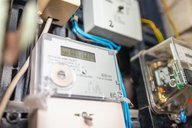 Das Bild zeigt einen digitalen Stromzähler, der an einer Wand montiert ist. Der Bildschirm zeigt die Verbrauchszahl „0003523.6 kWh“. Verschiedene elektrische Leitungen und weitere Geräte sind in der Nähe sichtbar.