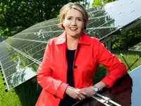 Eine lächelnde Frau in einem roten Mantel steht vor Solarpanelen in einem grünen Umfeld. Sie hat die Arme verschränkt und strahlt Zuversicht aus. Die Sonnenstrahlen reflektieren sich auf den Panels und betonen die umweltfreundliche Technologie.