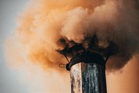 Eine rauchende Schornsteinöffnung wird von dichten, orangefarbenen Rauchwolken umschlossen. Der Rauch steigt in die Luft und deutet auf mögliche Umweltverschmutzung hin. Der Schornstein selbst ist aus Metall und hat eine abgenutzte Oberfläche.