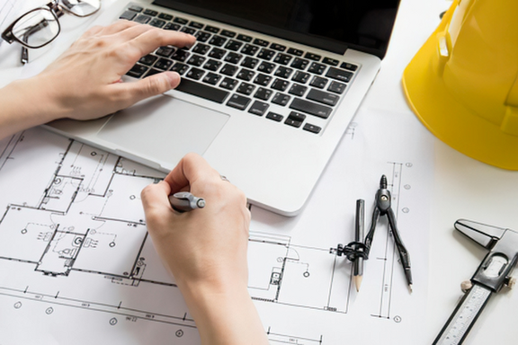 Eine Hand schreibt mit einem Stift auf Bauplänen, während die andere Hand auf einem Laptop tippt. Neben den Plänen liegen ein Zirkel, ein Bleistift und ein Maßband. Im Hintergrund steht ein gelber Bauhelm und eine Brille.