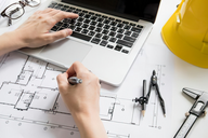 Eine Hand schreibt mit einem Stift auf Bauplänen, während die andere Hand auf einem Laptop tippt. Neben den Plänen liegen ein Zirkel, ein Bleistift und ein Maßband. Im Hintergrund steht ein gelber Bauhelm und eine Brille.