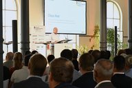Ein Redner steht an einem Podium und spricht vor einem Publikum in einem hellen Raum mit großen Fenstern. Auf dem Bildschirm und im Hintergrund sind Präsentationsmaterialien zu sehen. Das Publikum, bestehend aus mehreren Personen, hört aufmerksam zu.