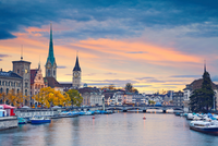 Ein Panoramablick auf Zürich bei Sonnenuntergang. Die Skyline zeigt mehrere markante Gebäude, darunter Kirchtürme und historische Fassaden. Der Zürichsee ist im Vordergrund sichtbar, während Boote am Ufer liegen. Der Himmel wird von warmen Farben des Sonnenuntergangs erleuchtet.