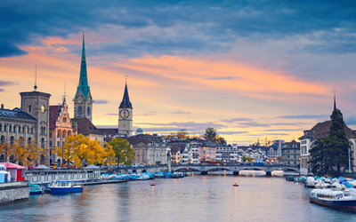 Ein Panoramablick auf Zürich bei Sonnenuntergang. Die Skyline zeigt mehrere markante Gebäude, darunter Kirchtürme und historische Fassaden. Der Zürichsee ist im Vordergrund sichtbar, während Boote am Ufer liegen. Der Himmel wird von warmen Farben des Sonnenuntergangs erleuchtet.