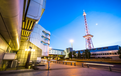 Eine moderne Architekturansicht mit einem futuristischen Gebäude im Vordergrund und einem hohen Sendeturm im Hintergrund. Der Himmel ist in sanften Blau- und Lilatönen gefärbt, während die Szene in den frühen Morgenstunden oder am Abend aufgenommen wurde.