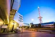 Eine moderne Architekturansicht mit einem futuristischen Gebäude im Vordergrund und einem hohen Sendeturm im Hintergrund. Der Himmel ist in sanften Blau- und Lilatönen gefärbt, während die Szene in den frühen Morgenstunden oder am Abend aufgenommen wurde.
