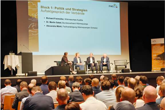 Eine Podiumsdiskussion mit vier Teilnehmern auf der Bühne, die über Politik und Strategien im Bereich Wärmepumpen sprechen. Im Hintergrund ist eine Präsentation an der Wand zu sehen, die den Titel „Block 1: Politik und Strategien“ trägt. Das Publikum sitzt vor der Bühne.