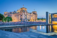 Das Bild zeigt den Reichstag in Berlin bei Dämmerung, beleuchtet und umgeben von Bäumen. Am Wasser sitzen viele Menschen, die auf das Gebäude blicken. Im Vordergrund verläuft eine moderne Treppe, die zu einem Wasserbecken führt.