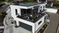 Das Bild zeigt ein modernes, zweigeschossiges Haus mit großen Fenstern und einem Balkon. Auf dem Balkon befinden sich Möbel und Pflanzen. Die Umgebung ist gepflegt mit einer Pflastersteinfahrbahn und kleinen Pflanzenbeeten.
