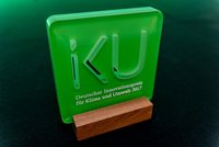 Die grüne Auszeichnung zeigt die Buchstaben "iKU" und trägt den Text "Deutscher Innovationspreis für Klima und Umwelt 2017". Der Preis steht auf einem Holzsockel und symbolisiert innovative Leistungen im Bereich Klimaschutz und Umwelt.