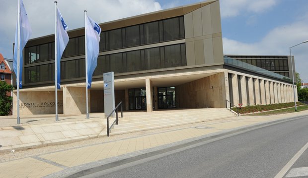 Modernes Gebäude der Industrie- und Handelskammer mit dem Namen "Ludwig-Bölkow". Die Fassade ist aus hellen Materialien und hat große Fenster. Vor dem Eingang stehen mehrere Fahnen und es gibt eine Treppe, die zu einem überdachten Eingangsbereich führt.