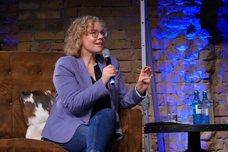 Eine Frau mit lockigem, blonden Haar und Brille sitzt auf einem Sofa und spricht in ein Mikrofon. Sie trägt einen blauen Blazer und Jeans. Auf einem Tisch neben ihr stehen Flaschen Wasser und ein Glas. Der Hintergrund hat eine strukturierte Mauer mit blauer Beleuchtung.