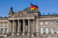 Das Bild zeigt die Fassade des Reichstagsgebäudes in Berlin mit der Aufschrift "Dem Deutschen Volke". Im Vordergrund weht eine Flagge Deutschlands, während im Hintergrund der charakteristische Kuppelbau sichtbar ist. Der Himmel ist klar und blau.
