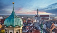 Blick auf die Stadt, mit einer charakteristischen grünen Kuppel im Vordergrund. Im Hintergrund erhebt sich ein hoher Kirchturm mit einem markanten roten Dach, umgeben von historischen Gebäuden in verschiedenen Farbtönen. Der Himmel zeigt sanfte, pastellfarbene Wolken bei Sonnenuntergang.