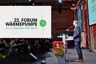 Ein Redner steht an einem Podium während des 23. Forums Wärmepumpe in Berlin. Im Hintergrund sind zahlreiche Zuschauer zu sehen. Auf der Leinwand ist das Event-Logo mit Datum und Ort eingeblendet. Frische Blumen stehen auf dem Tisch.