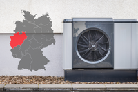 Eine Luftwärmepumpe steht an einer Wand, neben einer grauen Karte von Deutschland, die eine Region im Westen rot markiert. Das Bild zeigt die Kombination aus Heiz- und Kühltechnik und geografischer Lage.
