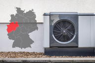 Eine Luftwärmepumpe steht an einer Wand, neben einer grauen Karte von Deutschland, die eine Region im Westen rot markiert. Das Bild zeigt die Kombination aus Heiz- und Kühltechnik und geografischer Lage.